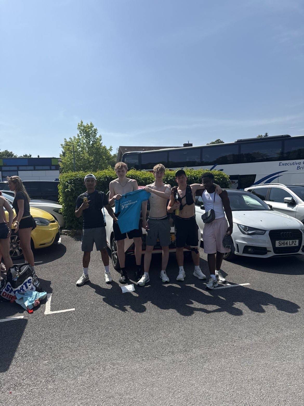 Group of six young friends posing in a sunny parking lot with cars and a bus in the background.