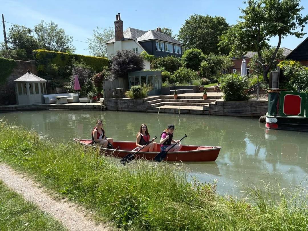 Three people in a red canoe paddling along a calm canal beside a landscaped garden and white gazebo.