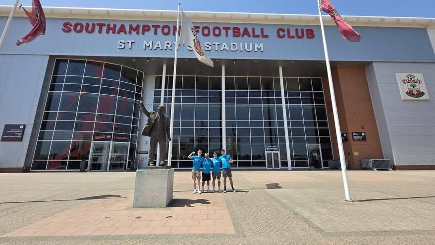 Front of Southampton FC's St Mary's Stadium with a statue in the foreground and four fans posing in blue shirts