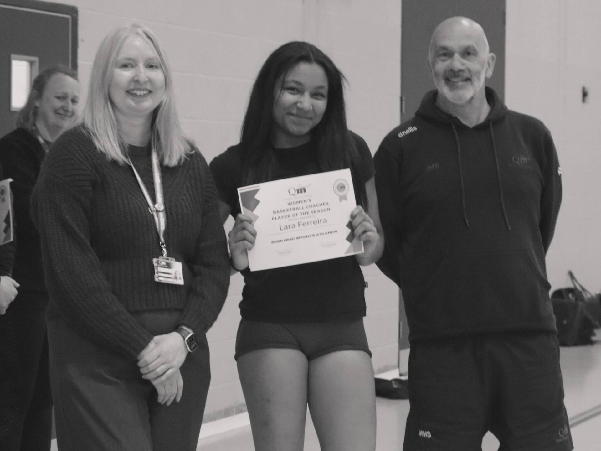 Group of four people posing in a gym; the central woman holds a certificate and smiles, flanked by two teammates and a man in a hoodie, all smiling.