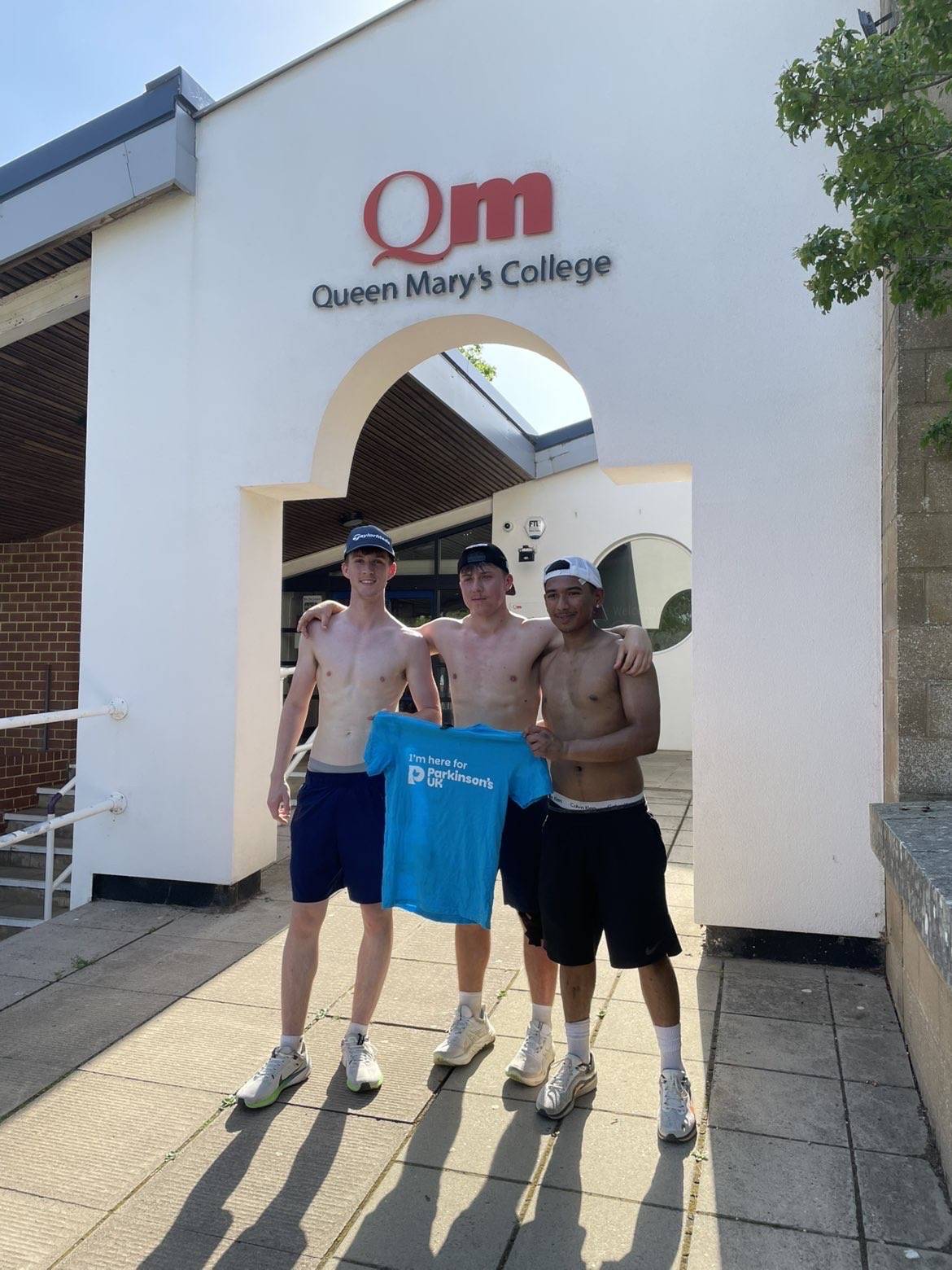 Three shirtless young men pose outside Queen Mary’s College, holding a blue Parkinson’s UK charity shirt in front of the QM entrance.