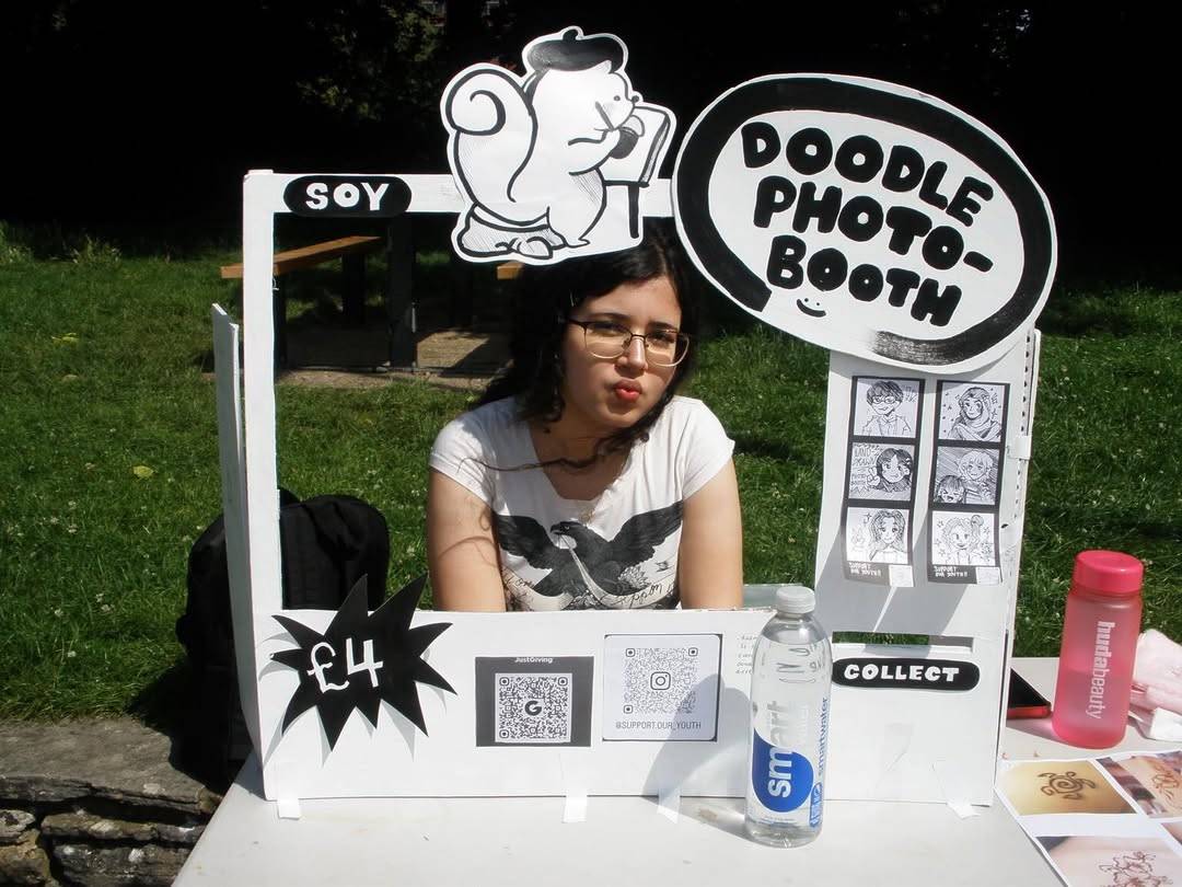 Girl with glasses sits at a DIY photo booth in a park; sign reads Doodle Photo-Booth, price £4 visible on a starburst shape.