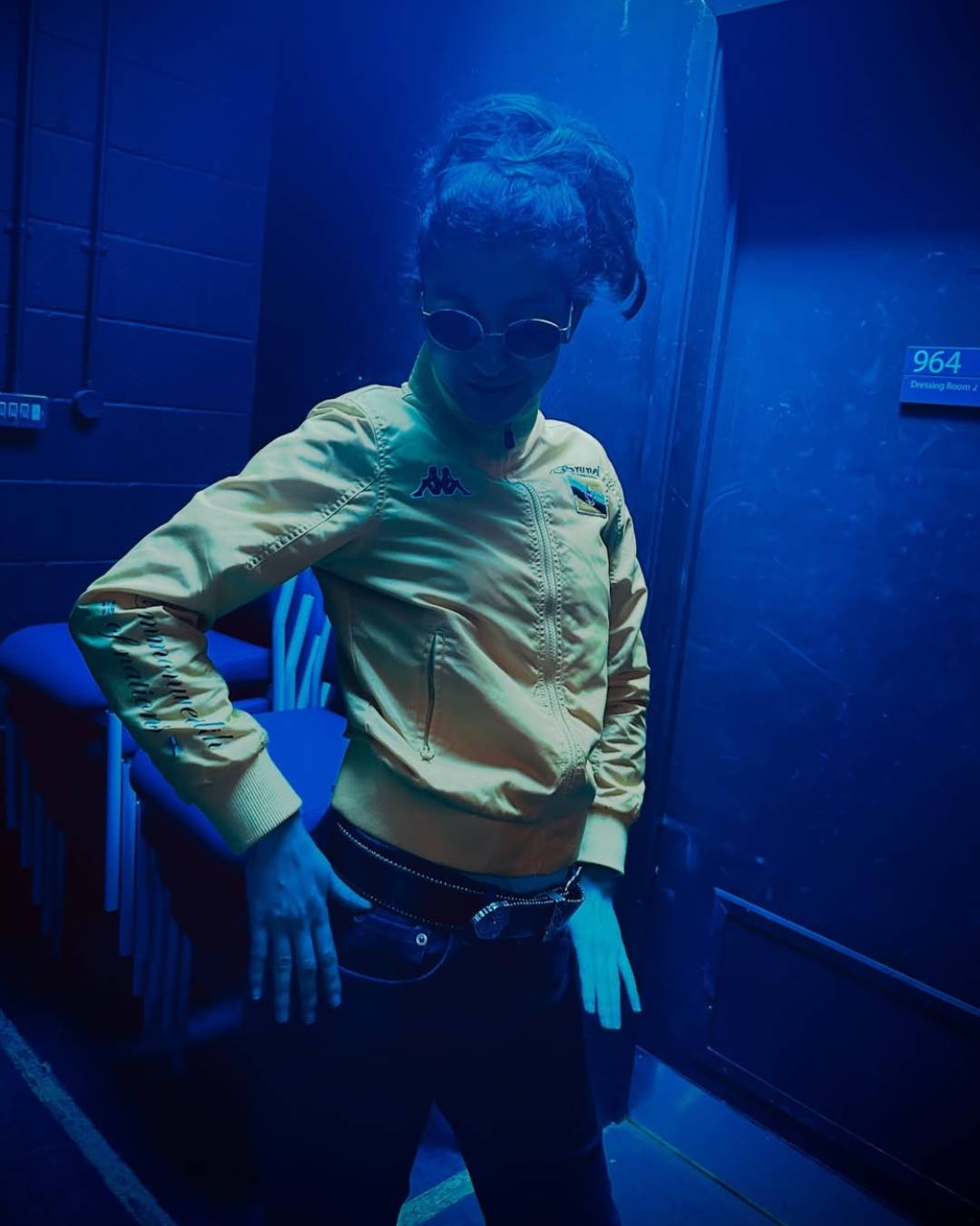 Person posing in a yellow jacket and sunglasses in a blue-lit backstage area, hands on hips.