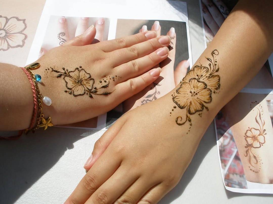 Two hands with intricate floral henna designs on the back of the left hand and the right forearm, bracelets visible.
