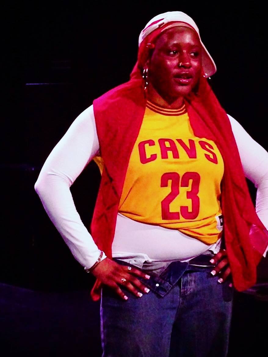 Person on a dark stage wearing a yellow Cavaliers jersey with number 23, a red sleeveless jacket, white long sleeves, jeans, and a head wrap, posing with hands on hips.