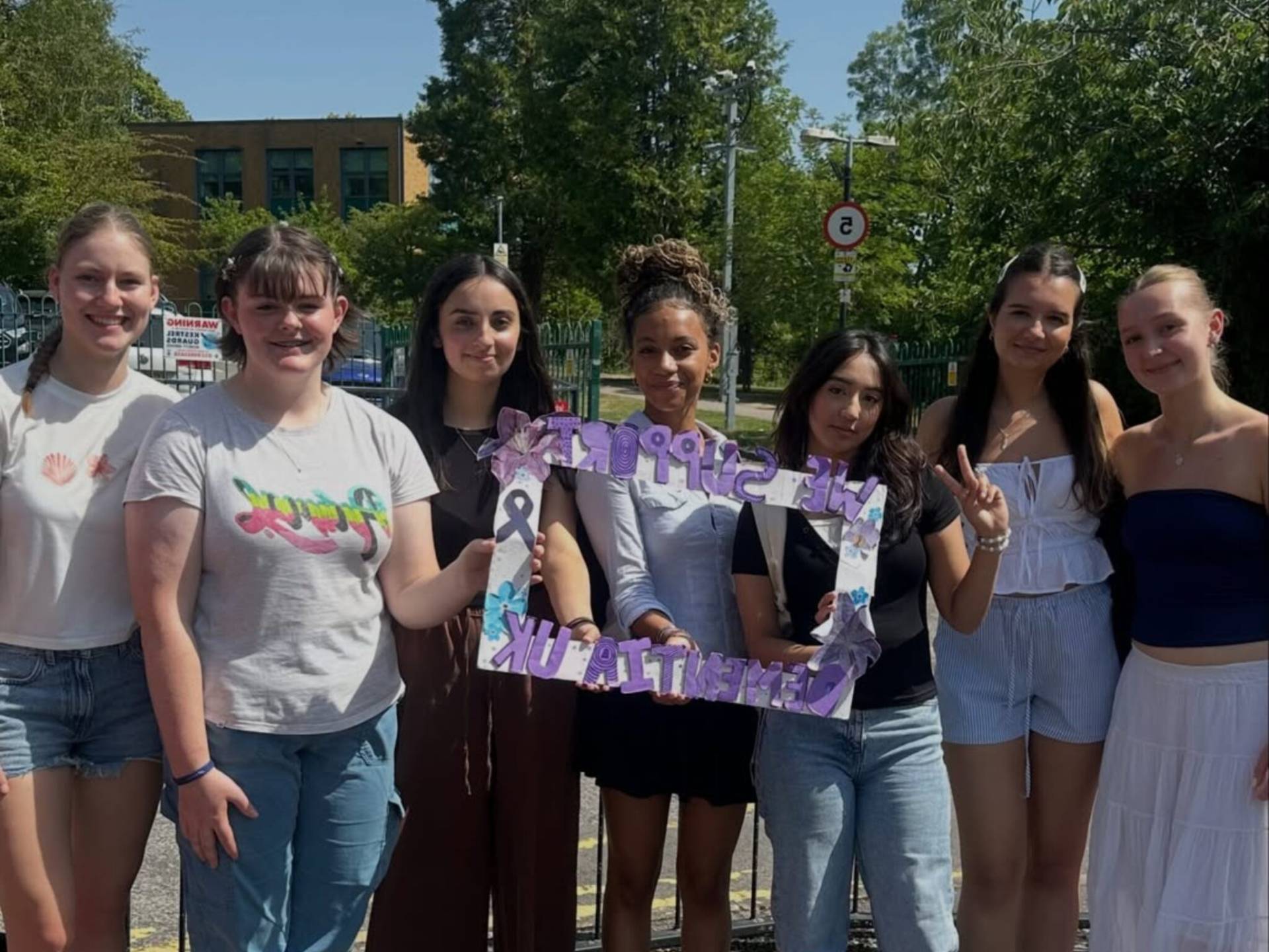 Group of seven young people outdoors, smiling, standing behind a metal fence and holding a purple banner.