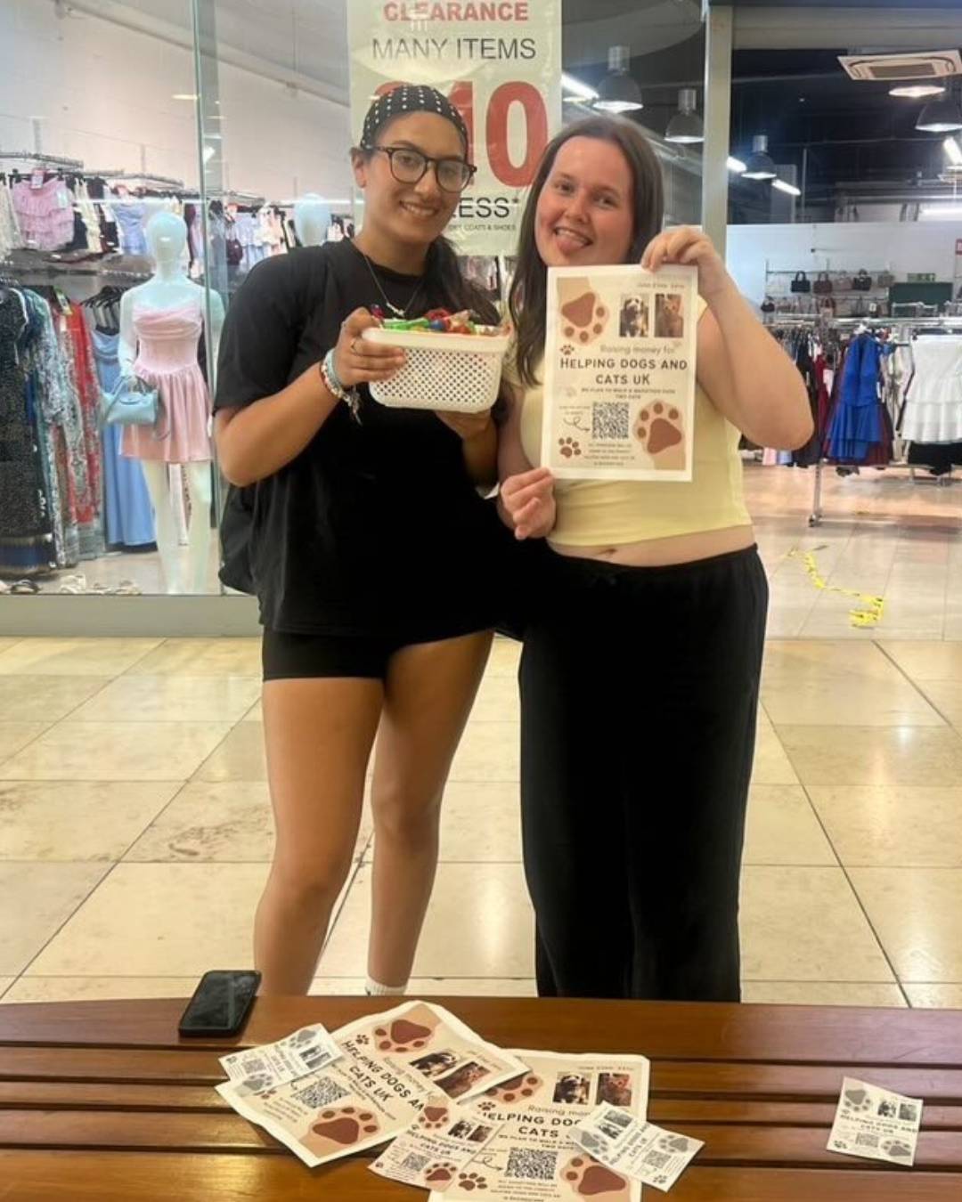 Two smiling women in a clothing store display a flyer for Helping Dogs and Cats UK, one holding a basket of candy and the other the flyer; flyers on a bench in front.